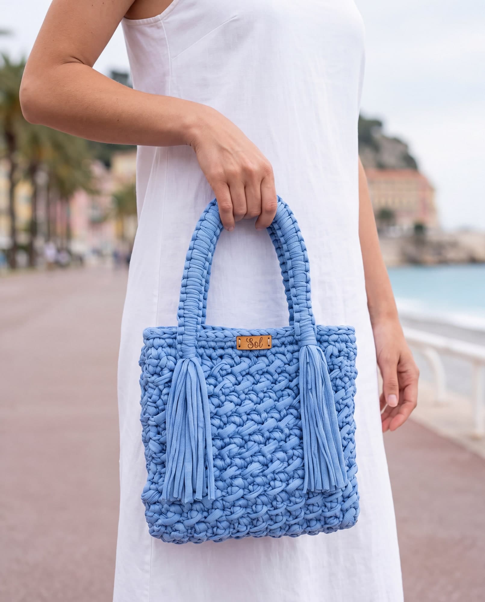 Mulher de vestido branco segurando a bolsa Nice azul-céu pelas alças, num calçadão à beira-mar, imagem ilustrativa