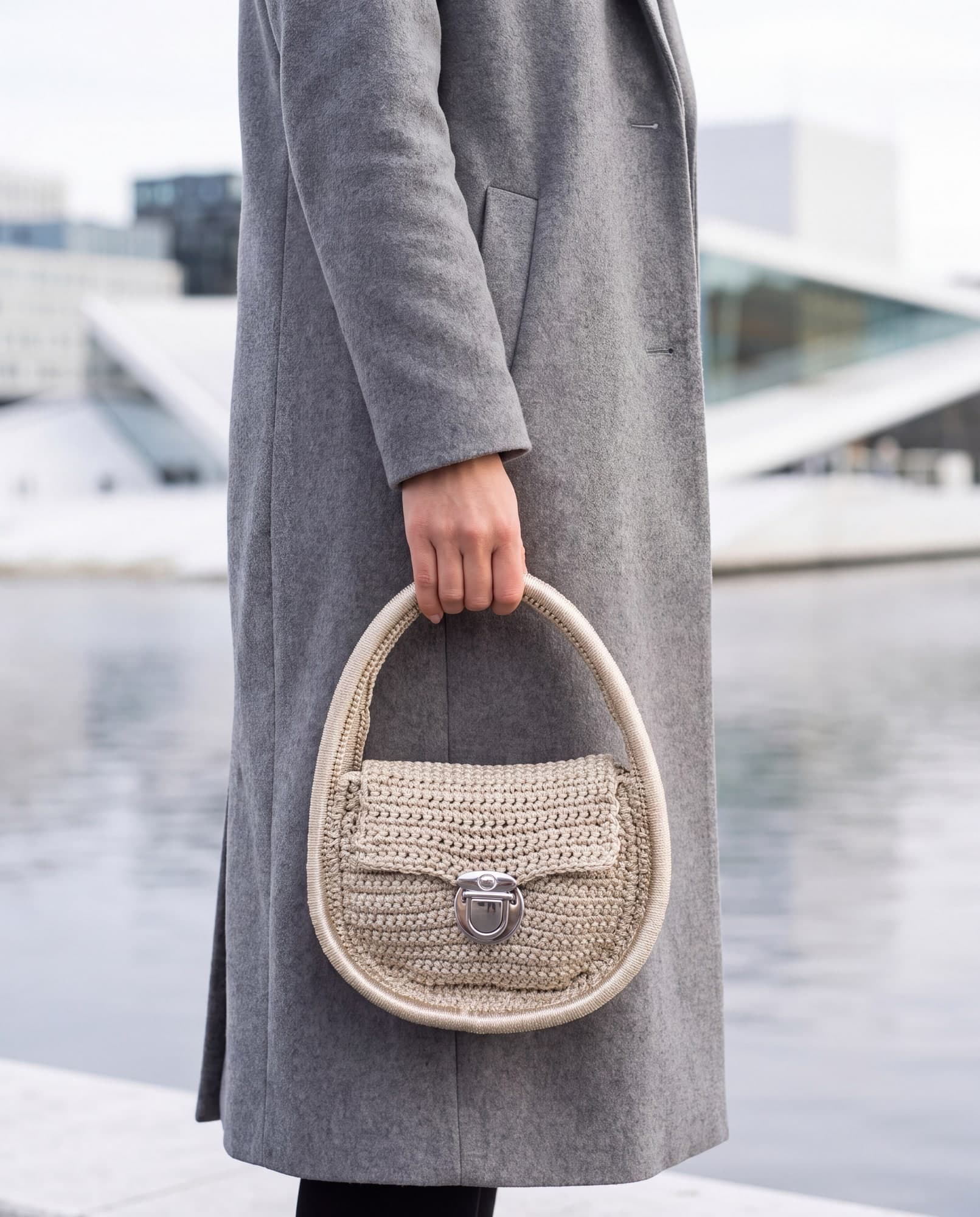 Mulher de casaco cinza segurando a bolsa Oslo pérola pela alça circular, à beira d'água, imagem ilustrativa